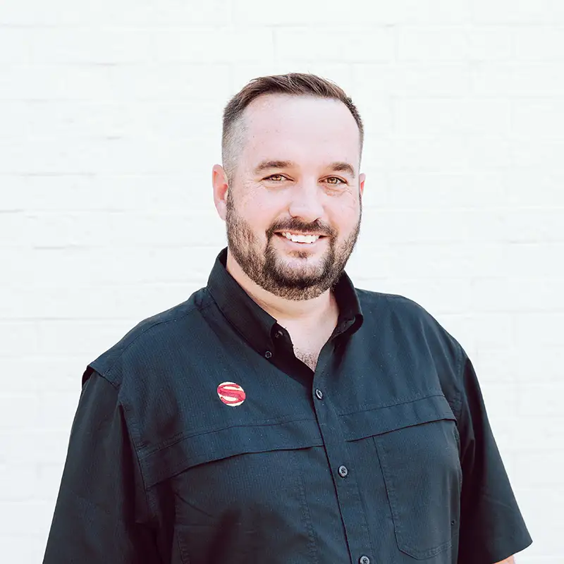 A man with short brown hair and a beard smiles at the camera. He is wearing a black button-up shirt with a red and white circular pin, standing in front of a white brick wall.