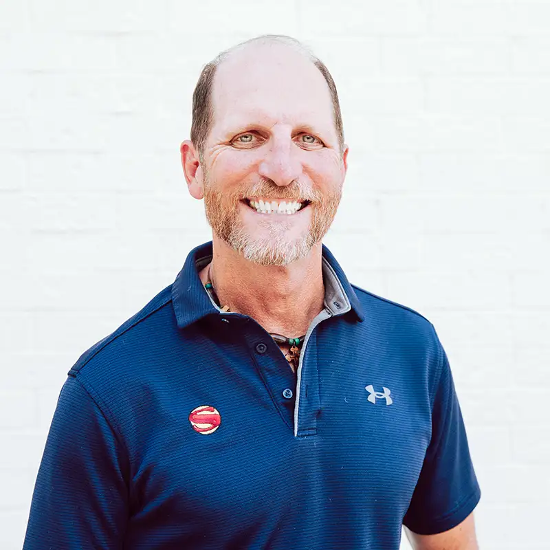 A smiling man with short hair and a beard wearing a navy blue Under Armour polo shirt stands in front of a white brick wall. He has a round red and white pin on his shirt.